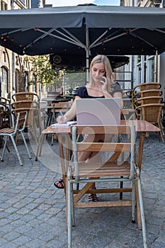 Attractive young blonde business woman using her laptop while calling with her smartphone, sitting outdoors in a