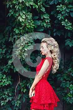 Attractive young blonde girl in red dress posing near a wild vineyard.