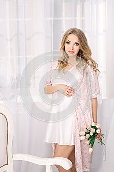 Attractive young blonde pregnant woman posing in white nightgown. Happy glamour girl at home.