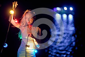 Attractive young blonde woman in summer dress holding colorful garland lamps at night. Water reflecion in background. Festive,