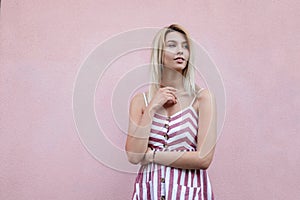 Attractive young blonde woman in a summer stylish striped sundress rests near a pink vintage wall in the city. Beautiful cute girl