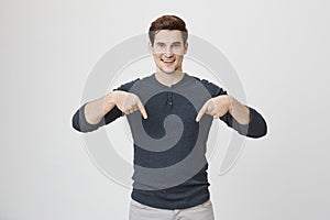 Attractive young boy with trendy haircut smiling and pointing down with both index fingers, standing over gray