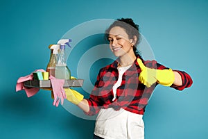 Attractive curly woman wearing yellow rubber gloves holds cleaning supplies in one hand and shows a thumb up to camera standing on