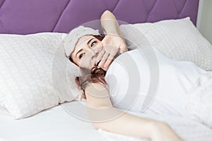 An attractive young brunette girl with brown hair wakes up and sips while yawning in her bed in a sleep mask