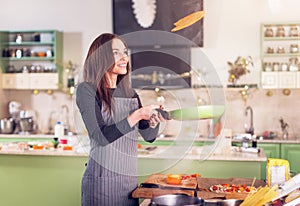 Attractive young brunette housewife in casual clothes and apron tossing pancakes on pan in the kitchen