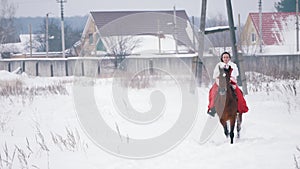 Attractive young brunette in a red dress galloping fast on a horse through the snow in the winter