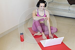 Attractive young brunette takes a break from exercises while sitting on a gym Mat, drinking water, listening to music with