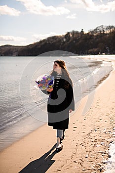 Attractive young brunette woman in black coat holding bright flower arragement standing by seaside