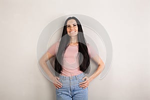 Attractive young brunette woman with long hair is standing by the grey background looking straight and smiling with arms