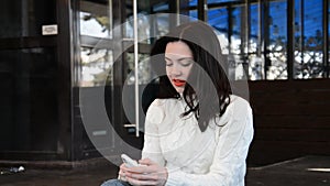 Attractive young brunette woman portrait in a white springe coat with red lips in the city tapping on mobile phone