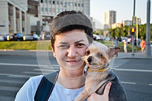 Attractive young brunette woman with short hair holds in her arms small cute Yorkshire terrier dog on background of city street.