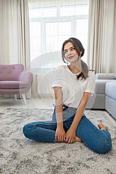 Attractive young brunette woman wearing jeans and white t-shirt sitting on the floor in bright livingroom