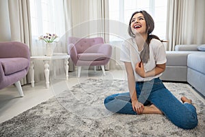 Attractive young brunette woman wearing jeans and white t-shirt sitting on the floor in bright livingroom and bursting