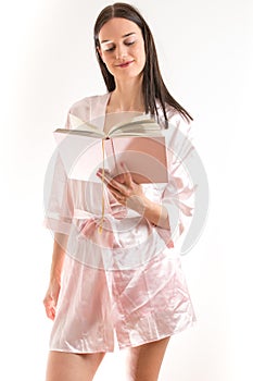 Attractive young brunette woman wearing a pink nightgown, reading a book with pink covers while standing