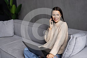 Attractive young business woman relaxing on a leather couch at home, working on laptop computer, talking on mobile phone