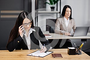 Attractive young business woman wearing jacket talking on mobile phone while sitting on a desk and using notebook near her