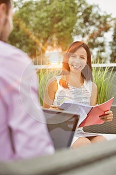 Attractive young business woman holding a file and smiling whilst she has a meeting outside wth a colleague who is working on his