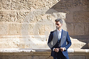 Attractive young businessman with beard and suit, posing next to a stone wall. Concept beauty, fashion, success, achiever, trend