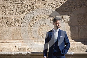 Attractive young businessman with beard and suit, posing next to a stone wall. Concept beauty, fashion, success, achiever, trend
