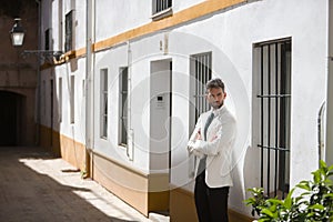 Attractive young businessman with beard, suit and tie, posing with arms crossed leaning against a sunny white wall. Concept beauty