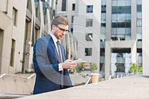 attractive young businessman in stylish suit with paper cup of coffee using smartphone