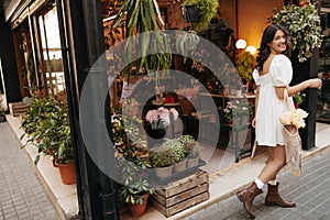 Attractive young caucasian girl holding string bag with bouquet near flower shop.