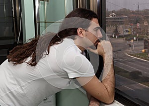 Attractive young caucasian guy with long hair and beard is standing bored on balcony due to Corona virus.