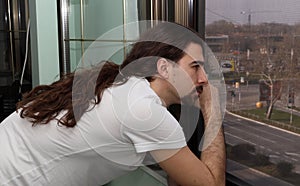 Attractive young caucasian guy with long hair and beard is standing bored on balcony due to Corona virus.