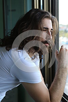 Attractive young caucasian guy with long hair and beard is standing bored on balcony due to Corona virus.