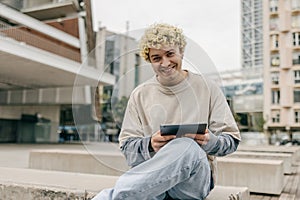 Attractive young caucasian man smiling in camera, using tablet sitting in yard of univercity.