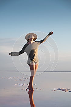 Attractive young Caucasian woman in a straw hat and long jacket walks balancing on a shallow salt lake at sunset