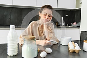 Attractive young cheerful girl baking at the kitchen, making dough, holding recipe book, having ideas