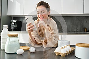 Attractive young cheerful girl baking at the kitchen, making dough, holding recipe book, having ideas