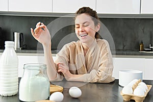 Attractive young cheerful girl baking at the kitchen, making dough, holding recipe book, having ideas