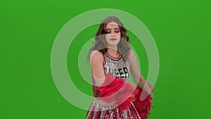 An attractive young cheerleader in uniform performs a jubilant dance with red pompoms in the studio on a green screen