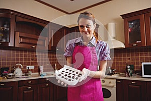 Attractive young confectioner wearing pink apron, standing in the middle of her home kitchen with a box of handmade chocolate