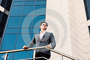 Attractive young confident businessman in classic suit is looking away, leaning on balcony balustrade, outside the office building