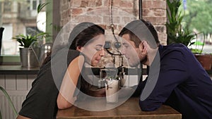 Attractive young couple in cafe sitting at the wooden table and sharing milkshake drinking it together using two straws