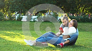 Attractive young couple at the city park sitting on the grass, hugging and looking at smartphone. Concept. Loving man
