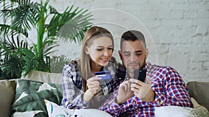 Attractive young couple with smartphone and credit card shopping on the internet sit on couch in living room at home