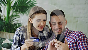 Attractive young couple with smartphone and credit card shopping on the internet sit on couch in living room at home