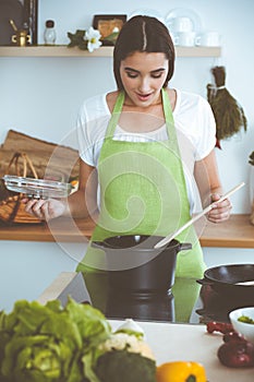 An attractive young dark-haired woman preparing soup by new keto recipe while standing and smiling in the kitchen