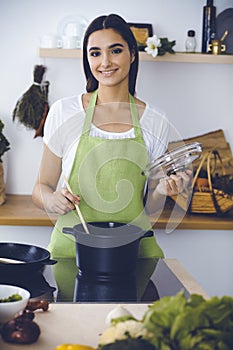 An attractive young dark-haired woman preparing soup by new keto recipe while standing and smiling in the kitchen