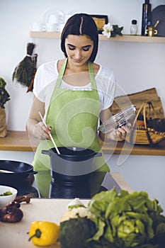 An attractive young dark-haired woman preparing soup by new keto recipe while standing and smiling in the kitchen