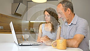 Attractive young daughter and senior father are using laptop sitting in modern kitchen in apartment. Woman is teaching