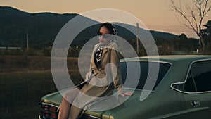 Attractive young elegant woman in a retro outfit sits on the hood of her car.