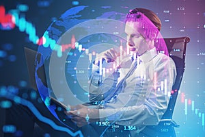 Attractive young european businessman sitting and looking at laptop with glowing downward candlestick forex chart on blurry