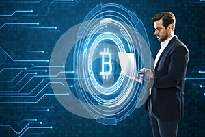 Attractive young european businessman using laptop with creative glowing crypto coins on dark blue background with circuit lines.
