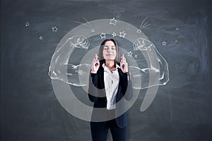 Attractive young european businesswoman with crossed fingers standing on chalkboard wall background with drawn muscle hands.