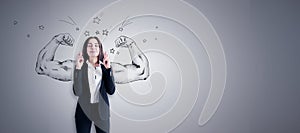 Attractive young european businesswoman with crossed fingers standing on concrete wall background with drawn muscle hands and mock